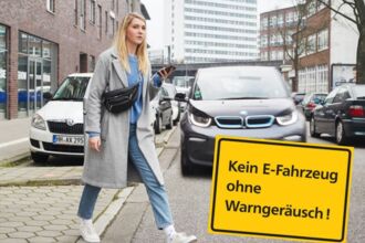 Eine junge blonde Frau mit grauem Mantel, Jeans und Smartphone in der Hand betritt eine Straße, auf der sich ihr ein Auto nähert. Ihr Blick ist dem Auto abgewandt. Im Bild ist ein gelbes Schild mit schwarzer Aufschrift: Kein E-Fahrzeug ohne Warngeräusch!