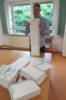 Ein Mann, mitte dreißig, steht und hält eine Wahlschablone in den Händen, vor ihm liegen stapelweise Wahlschablonen, Cd´s und Briefumschläge